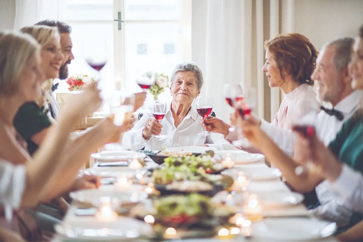 Group of people toasting with wine glasses around a dining table set with food. An elderly woman sits at the center.