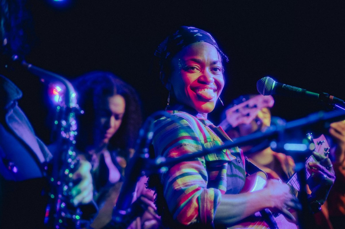 Musician smiles while playing guitar on stage, colorful lights illuminate her and band members in the background.